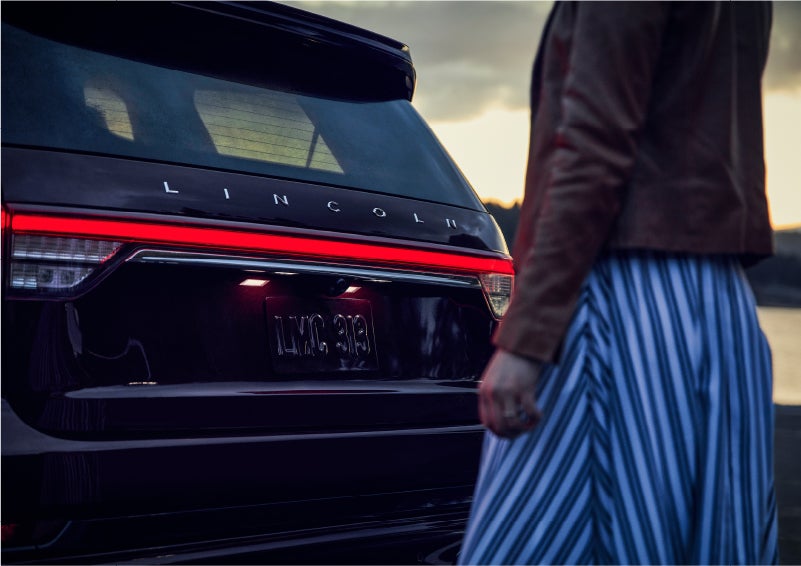 A person is shown near the rear of a 2023 Lincoln Aviator® SUV as the Lincoln Embrace illuminates the rear lights | Spitzer Lincoln DuBois in Du Bois PA