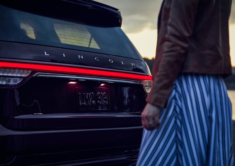 A person is shown near the rear of a 2024 Lincoln Aviator® SUV as the Lincoln Embrace illuminates the rear lights | Spitzer Lincoln DuBois in Du Bois PA
