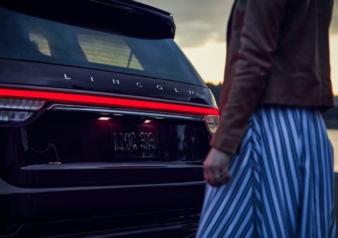 A person is shown near the rear of a 2024 Lincoln Aviator® SUV as the Lincoln Embrace illuminates the rear lights | Spitzer Lincoln DuBois in Du Bois PA